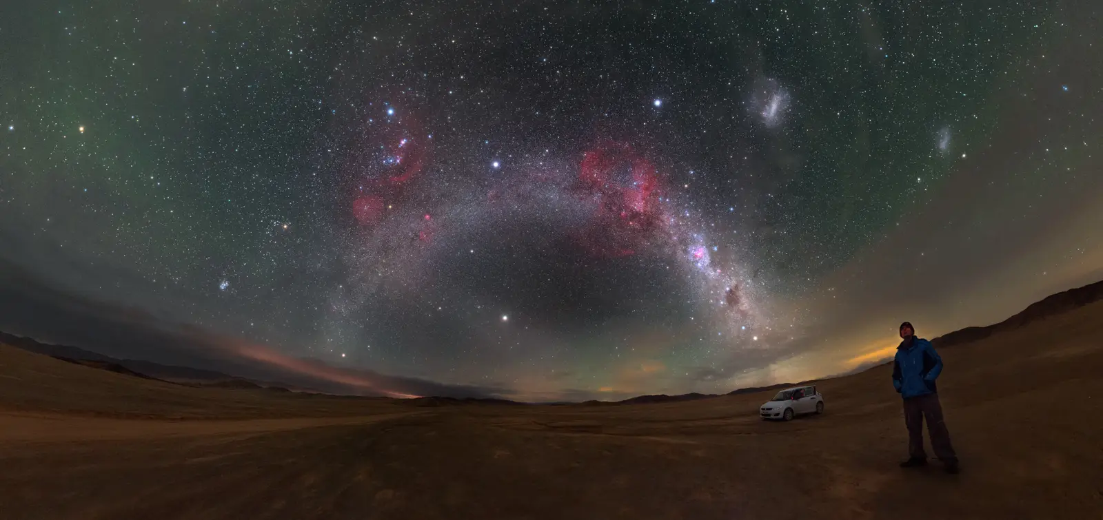A deep wide-angle night-sky panorama over the Atacama Desert: the arch of the Milky Way with the pink half-ring of Barnard’s Loop, and the Small and Large Magellanic Clouds as two pale diffuse patches at upper right.