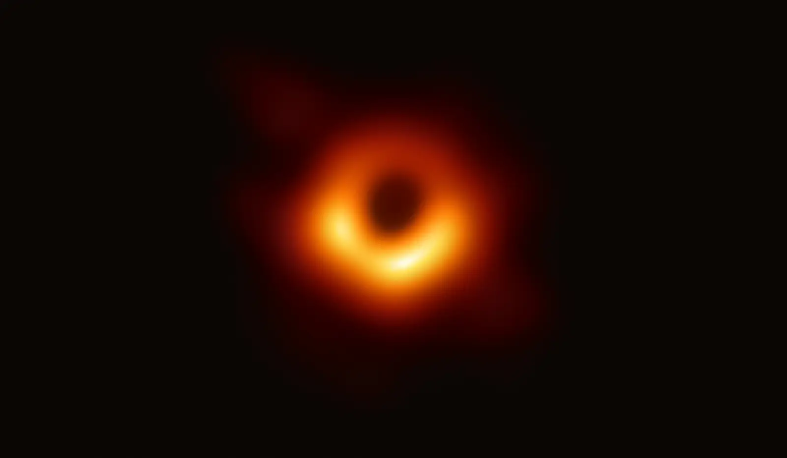 The first image of a black hole: a fuzzy orange ring of light, brighter along its lower edge, surrounding a dark central shadow.