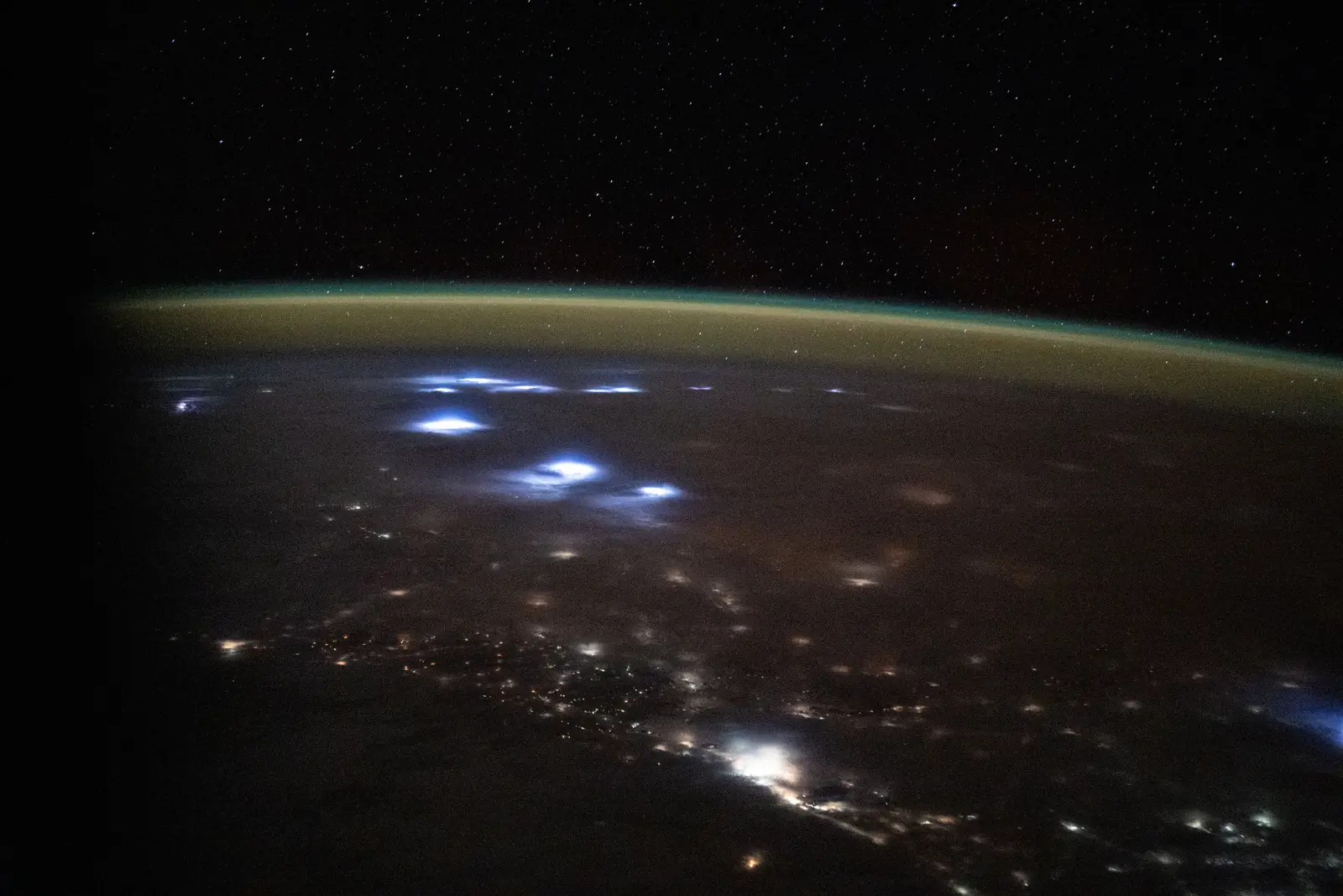 Night-side Earth photographed from the International Space Station: the scattered orange city lights of western Africa, blue flares of lightning inside cloud tops, a green-gold band of airglow along the limb, and stars in the black above it.