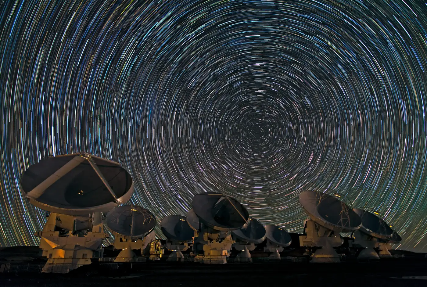 A long-exposure star-trail image over the ALMA antennas: every star drawn out into a concentric arc around the celestial pole.